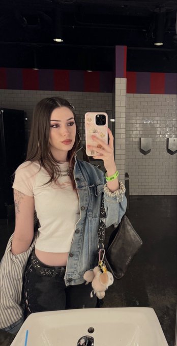 A young woman with long brown hair takes a mirror selfie in a bathroom, holding a phone with a pink floral case, wearing a white crop top and denim jacket with a small plush charm hanging from her bag. The image ties into the post's promotional context as a lifestyle/selfie-style post promoting the @Stake monthly $100,000 race and BTCsRewards leaderboard offer with code "gray."