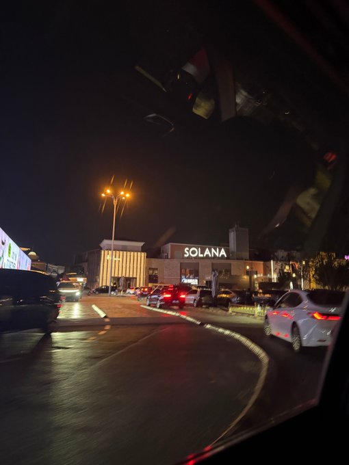 A nighttime street view taken from inside a car, showing a building with 'SOLANA' illuminated on its facade and traffic on the wet road.