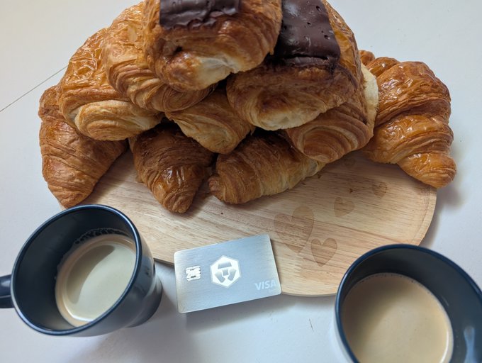 A wooden board holds a generous pile of golden croissants—one topped with a square of chocolate—flanked by two mugs of coffee, with a silver Crypto.com Visa card placed in front, perfectly illustrating a slow Sunday morning spent stacking $CRO cashback.