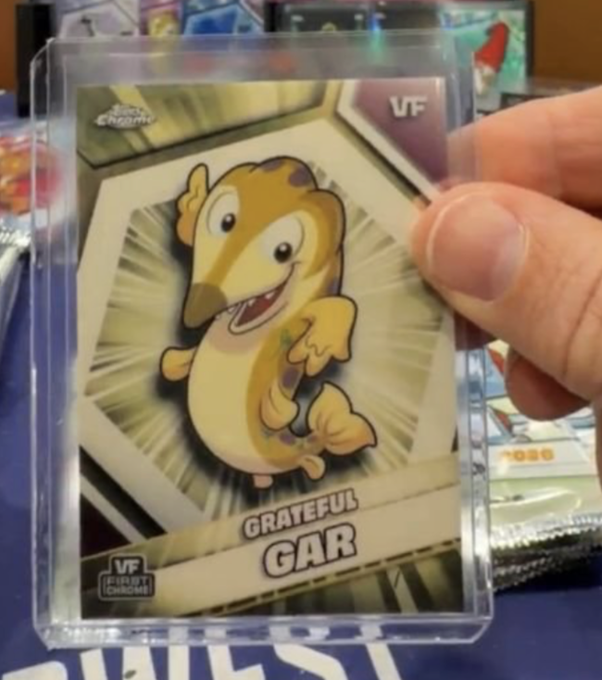 The image features a person holding up a small, cartoon-style card with the text "Grateful Gar" written on it. The card has a fish graphic of a goldfish with a yellow body and black fins, which appears to be pulled off by a force or explosion. The background is blurred, but there are other cards visible in the image as well. The post includes a reference to the card being from the "VeeFriendsCards" series on Twitter, indicating that it might be related to a social media platform or event.