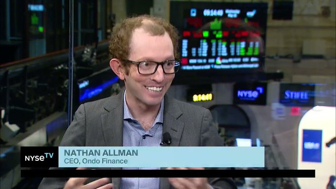 The image shows a man wearing glasses sitting at a desk with a computer monitor behind him. The screen displays a news segment about Nathan Allman's passing. The caption below the image reads, "Just In: @OndoFinance says its founder, Nathan Allman, has unexpectedly passed away."