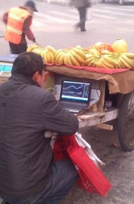Photograph of a street fruit vendor sitting on the ground using a laptop displaying stock/crypto charts, with bunches of bananas and apples displayed on a cart in front of him.
