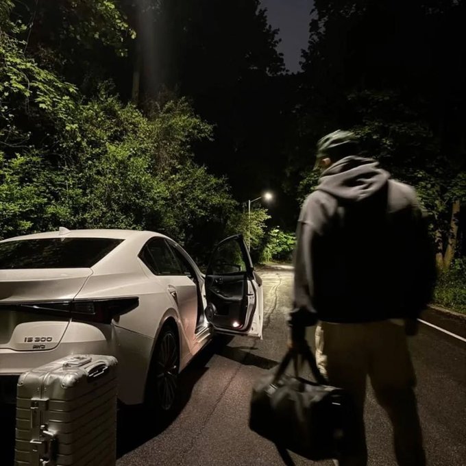 A nighttime scene of a person with a backpack and helmet carrying a silver suitcase toward a white Lexus IS 500 car with its door open on a tree-lined road.
