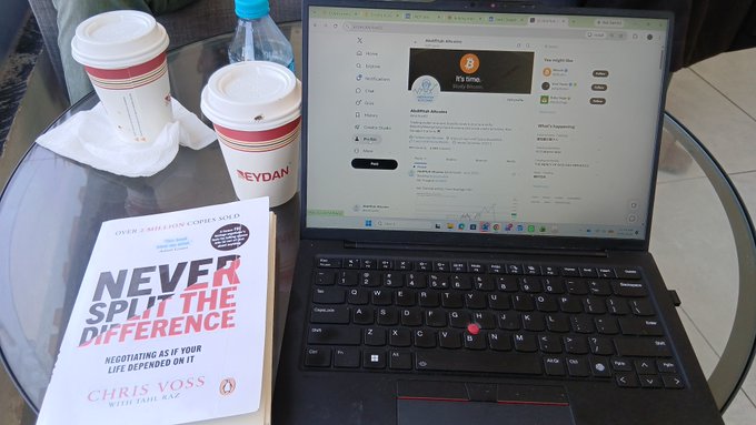 A laptop displaying an 'AutoFold Albums' webpage sits on a glass table alongside two 'MEYDAN' coffee cups and a book titled 'NEVER SPLIT THE DIFFERENCE' by Chris Voss.