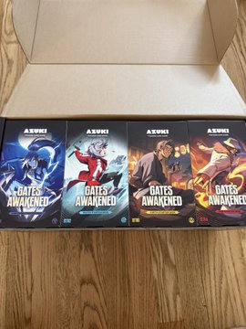 Four manga volumes of 'GATES AWAKENED' by A.SW1 are lined up in a cardboard box on a wooden floor, featuring colorful cover art.