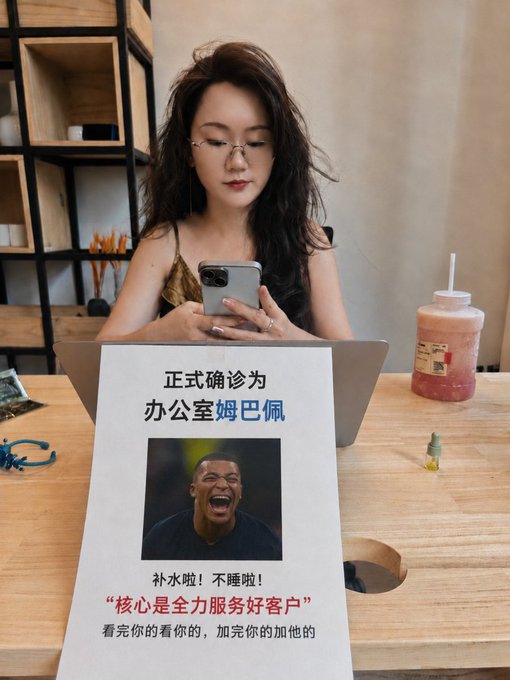 A young woman with glasses looking at her phone while holding a sign with Chinese text and an image of Kylian Mbappé laughing, poking fun at office burnout culture.