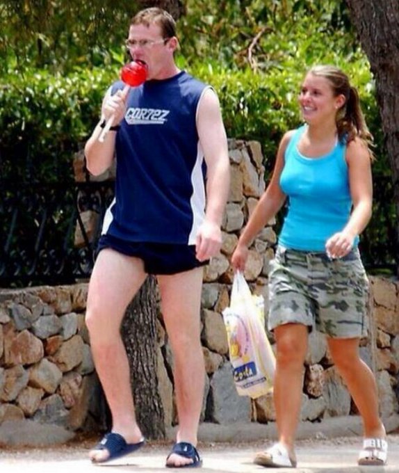 A man wearing shorts and glasses, holding a red drink, and a woman carrying a bag are walking together down a path near some trees. The couple appears excited about an upcoming event called EthConf, which they have been looking forward to attending.