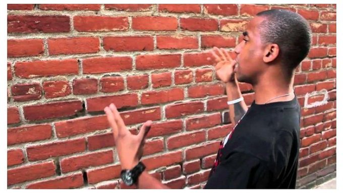 A young man in a black t-shirt and wristwatch stands in profile against a red brick wall, gesturing with his hands as if speaking or rapping.