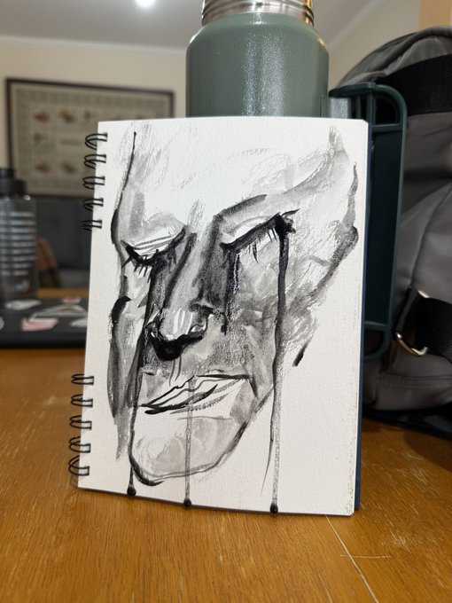 A black and white ink or charcoal drawing of a stylized face with closed eyes and dark dripping tears is rendered on a spiral-bound sketchbook resting on a wooden surface.
