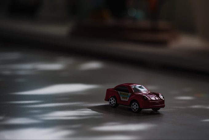A small red toy car (labeled 'Strawberry Drive') driving on a dark surface with light reflections, rendered in a cinematic, moody style with shallow depth of field.