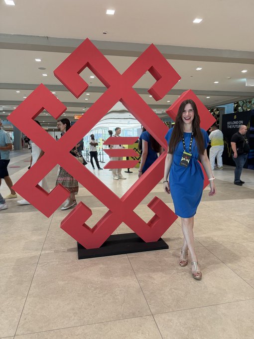 A smiling woman in a blue dress standing next to a large red geometric sculpture in what appears to be a conference or convention hall.