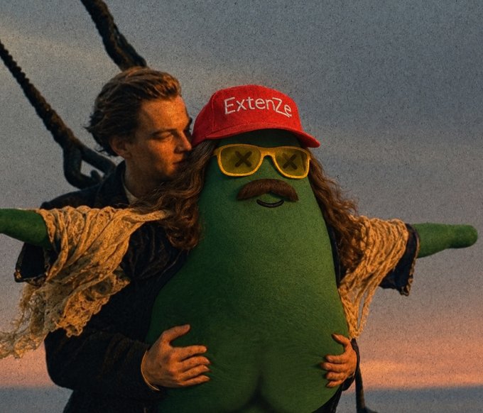 A person holding up a plush toy resembling a pickle character wearing a red 'ExtenZe' cap and yellow 'X' sunglasses, with ropes visible in the background.