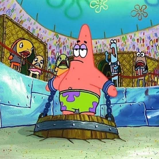 Patrick Star from SpongeBob SquarePants stands in an arena with a stoic expression, wearing green and purple shorts and chained to a wooden barrel with metal spikes.