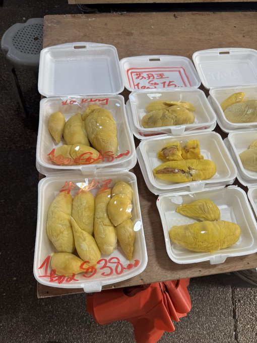 Several styrofoam containers on a wooden surface displaying fresh yellow durian fruit pieces, with red handwritten text showing prices like '1 ลูก $350.00'.