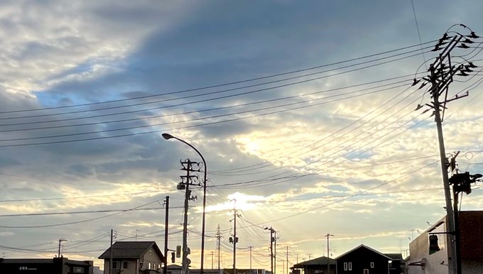 A suburban street scene at sunset with numerous power lines crisscrossing the sky, utility poles, street lamps, and houses silhouetted against dramatic clouds.