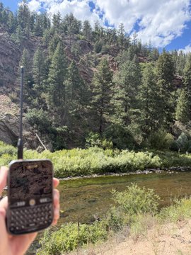 A hand holds a two-way radio in the foreground beside a clear river, dense pine trees, rocky hills, and a partly cloudy sky.