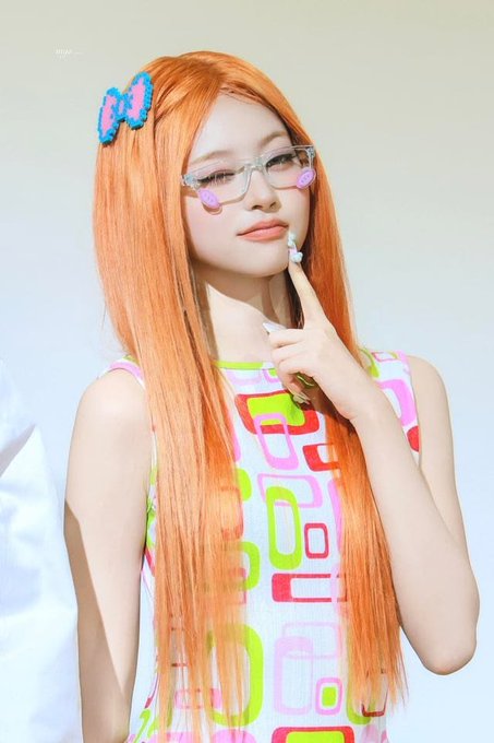 A young woman with long, vibrant orange hair poses with her finger thoughtfully touching her chin. She wears clear-framed glasses decorated with pink flower accents and a colorful hair clip featuring a blue and pink bow design. Her sleeveless top displays a playful geometric pattern of rounded rectangles in shades of pink, green, orange, and red on a white background.