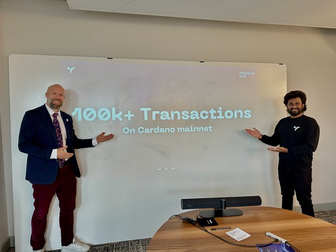 The image features two men standing next to each other, both wearing suits and ties, against a white background. They are positioned in front of a large screen displaying text related to 100,000 transactions on Cardano. The men appear excited about this achievement, possibly discussing their plans or strategies for the project.