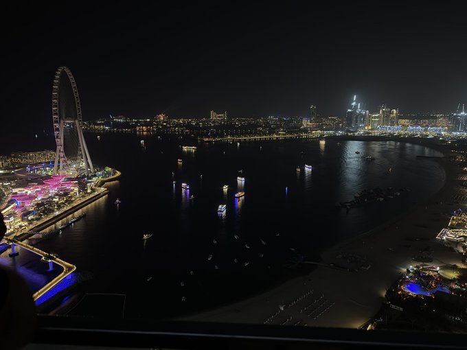 A sweeping nighttime aerial view captures the illuminated Ain Dubai observation wheel on the left and the glittering skyline of Dubai Marina across the bay, taken from a high-rise balcony in good night with boats dotting the water below.