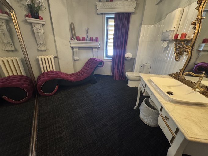 An ornate, baroque-style bathroom featuring a tufted purple velvet chaise lounge, a gold-framed mirror, a white sink, and a toilet against dark flooring.