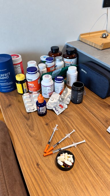 A wooden table displays an assortment of supplement bottles—including TMG, Perfect Amino, and Fish Oil—alongside syringes, pill packets, and a black case concealing injectables, illustrating a personalized and carefully curated stack of nootropics and supplements that required precision testing to develop.