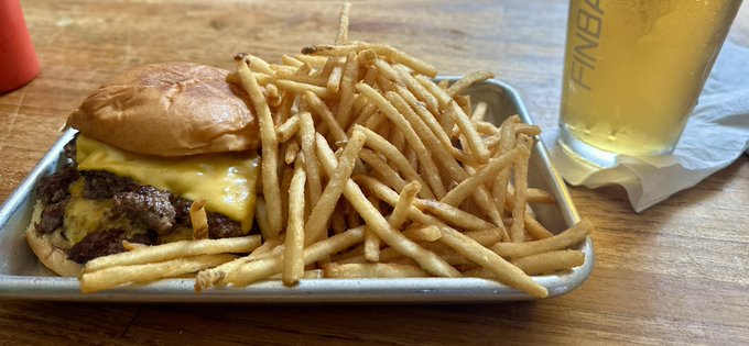 A double cheeseburger with melted cheese sits on a metal tray piled high with crispy shoestring fries, accompanied by a glass of FINBACK beer.