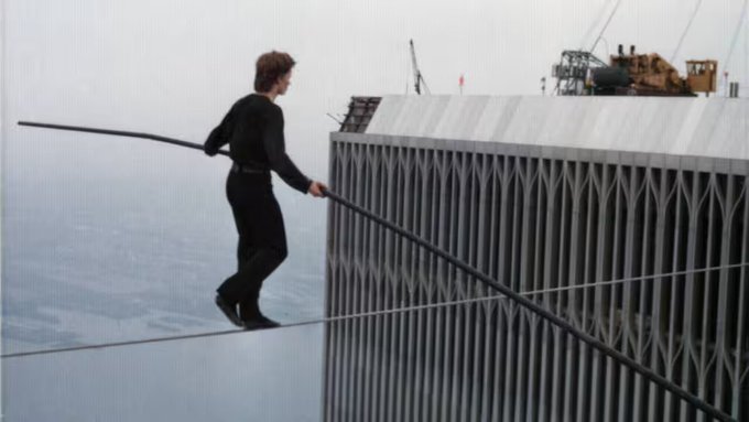A man in black clothing walks across a tightrope while balancing a long pole, performing high above a city with construction cranes visible in the background.