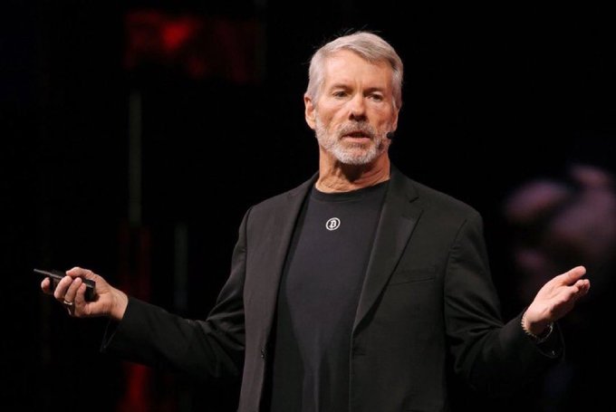 A man with gray hair and a beard, wearing a black suit jacket over a black t-shirt, is on stage mid-speech, holding a small device and gesturing with his hands. He appears to be a presenter at a conference or event, possibly addressing the recent news about a major figure selling 1,637 bitcoins, as referenced in the caption.