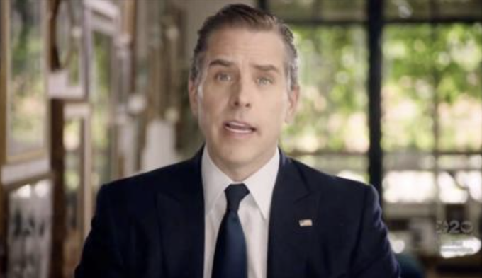 A man in a dark suit, white shirt, and navy tie addresses the camera indoors, with a small U.S. flag pin and a faint “20” watermark visible.
