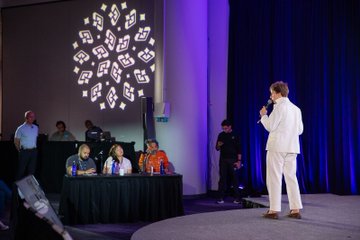 The image shows a stage with a woman standing behind a microphone, giving a speech or presentation. There are four people visible in the scene - one person sitting at a table, another person standing next to her, and two more individuals seated on chairs. The setting appears to be an event where speakers present their ideas, possibly related to cryptocurrency.