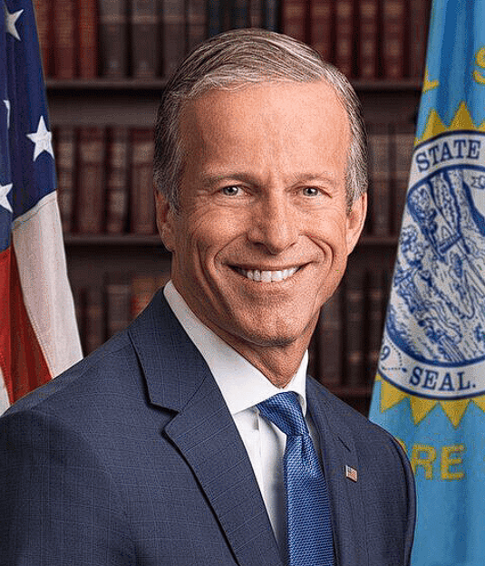 A man in a navy blue suit and blue tie smiles in an official portrait, positioned between an American flag and a blue state flag bearing the visible text "STATE SEAL." The image depicts US Senate Majority Leader John Thune, the official referenced in the post about the delayed CLARITY Act and postponed BTC crypto regulation vote.