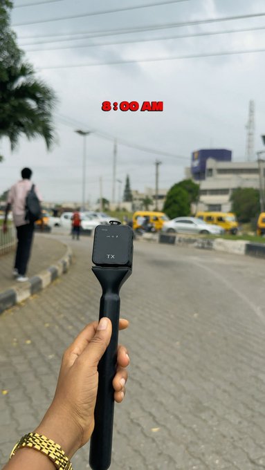A person holding a black handheld selfie stick or camera mount in front of a street scene with yellow buses, overlaid with the text '8:00 AM'.