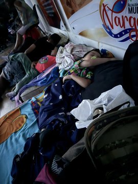 A woman lies on a blanket on the street outside a building with a "Vargas" sign, resting on bedding and surrounded by belongings after being displaced by earthquakes in Venezuela.