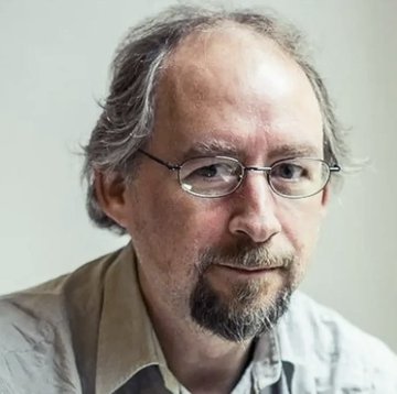 A close-up portrait shows a middle-aged man with graying hair, a beard, wire-rimmed glasses, and a light collared shirt, looking directly at the camera with a neutral expression. This image represents Adam Back, the cited expert, whose bullish quote accompanies the photo, lending authority to the claim about a potential U.S. Strategic Bitcoin Reserve driving Bitcoin to seven-figure valuations.