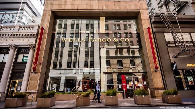 A street-level photograph captures the entrance to a Wells Fargo & Co. bank branch, featuring large glass windows, red banners, and pedestrians walking by on the city sidewalk.