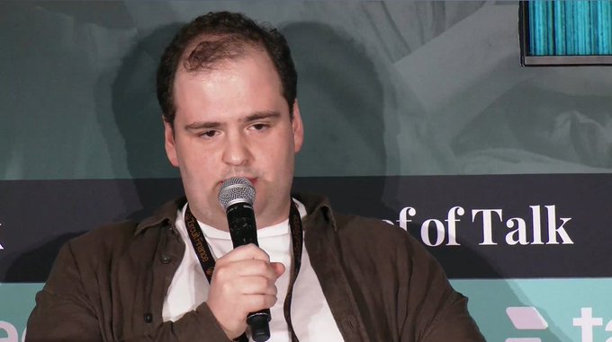 A man with short dark hair holding a microphone, speaking at an event with 'Talk' visible on a sign behind him.