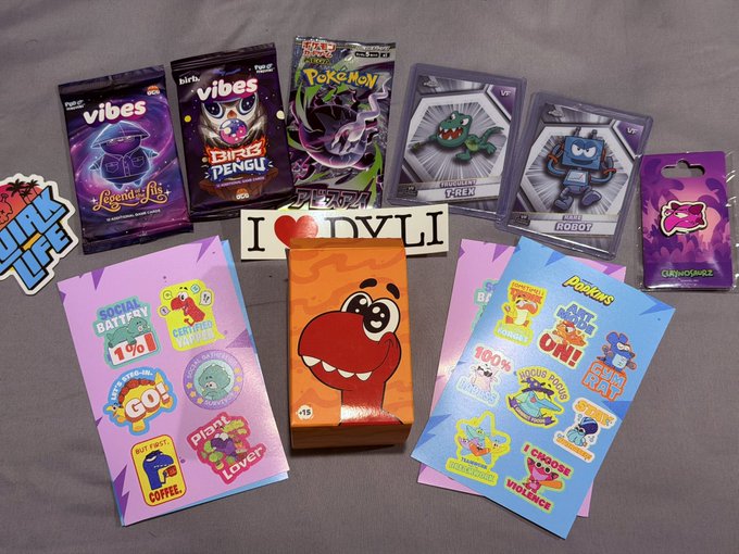 An assortment of merchandise is displayed on a gray surface, including 'VIBES' and 'BIRDS PENGU' cards, Pokemon collectibles, Robot figures, stickers, and packages featuring cartoon dinosaurs.