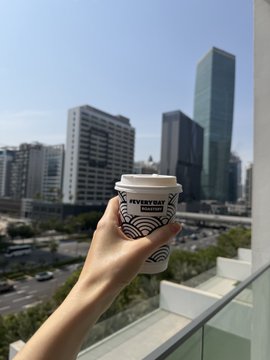 A hand holds a University of Tokyo coffee cup against a backdrop of Tokyo city buildings on a sunny day.