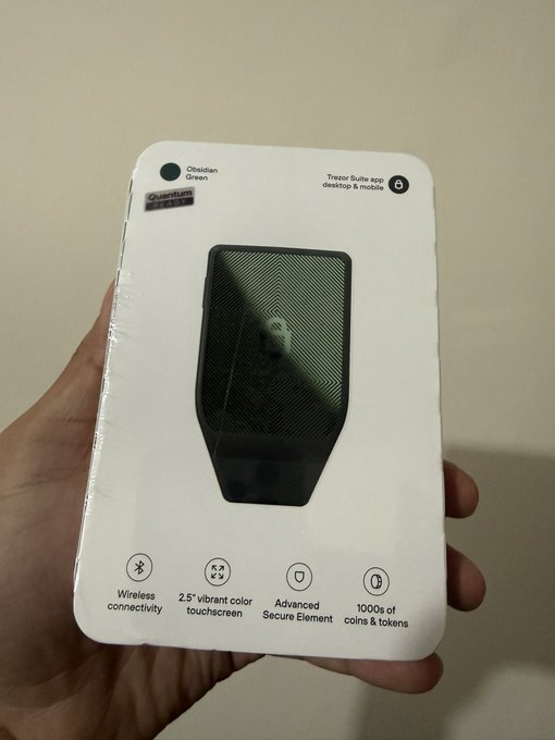 A person's hand holds the retail packaging for an Obsidian Green Trezor Safe 3 cryptocurrency hardware wallet, with the post noting its arrival alongside positive impressions of Tangem and SafePal alternatives. The box highlights features like wireless connectivity, a 2.5" vibrant touchscreen, and advanced secure element support.