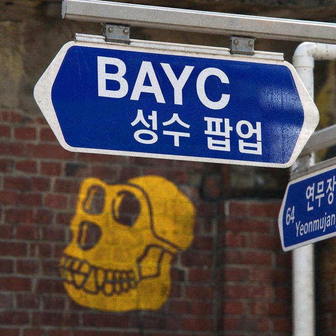 A blue street sign reading 'BAYC 성수 팝업' (BAYC Seongsu Popup) in English and Korean, with a yellow spray-painted skull mural visible on a brick wall behind.