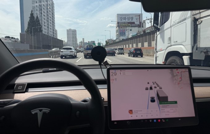A view from inside a Tesla shows the steering wheel with the brand's logo and the central touchscreen displaying an Autopilot visualization of surrounding traffic at 29 mph, while the car travels on a highway alongside a large truck and other vehicles toward a Korean cityscape. The image illustrates the poster's claim of using Autopilot software version SW 2026.14.6 on what they believe may be their last Tesla.