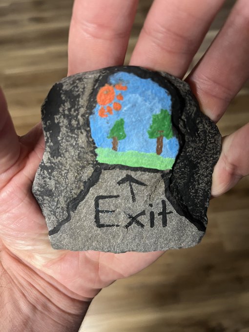 A hand holds a painted rock depicting a cave entrance with a blue sky, orange sun, green trees, and grass, with an arrow and the word 'Exit' written below.