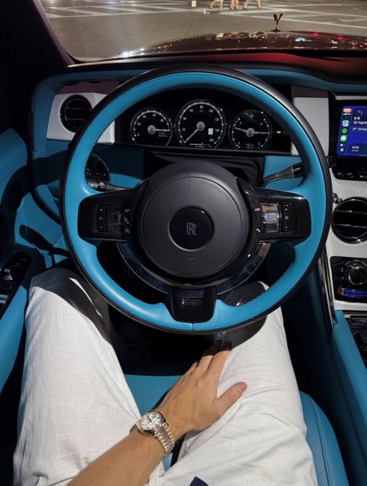 The image shows a person sitting in the driver's seat of a luxury Rolls-Royce, featuring a striking turquoise blue and black leather interior with the iconic "RR" logo on the steering wheel. The driver is wearing white pants and a watch, with the nighttime cityscape visible through the windshield. This opulent scene complements the post's crypto promotion ("1.5 $SOL Just hit the like and drop your wallets"), using the luxury car aesthetic to signal wealth and success typically associated with crypto gains.