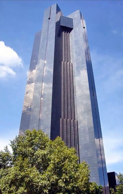 A tall, modern skyscraper with a reflective dark glass and bronze-colored facade, photographed from a low angle against a blue sky with white clouds and green trees at the base.