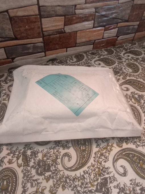 A white plastic package with a green label sits on a paisley-patterned fabric surface against a stone-tiled wall.