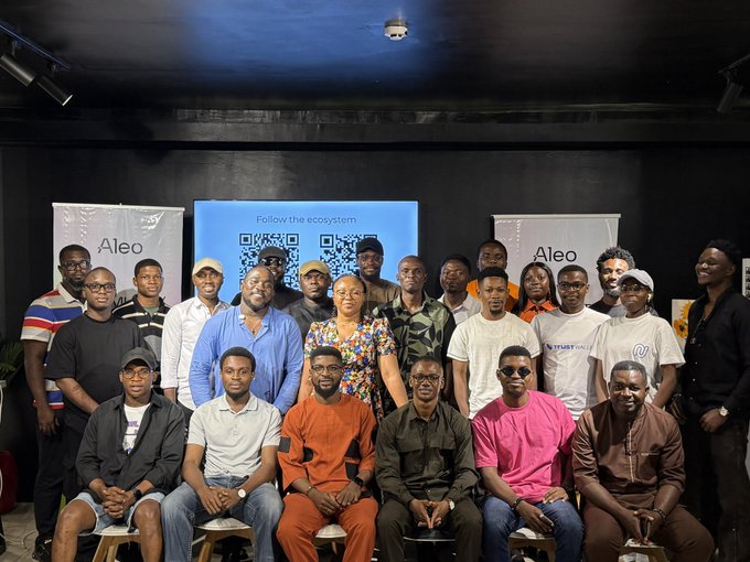 A group photo of approximately 25 people posing together at an Aleo event, with a banner in the background reading 'Follow the ecosystem' and a QR code.