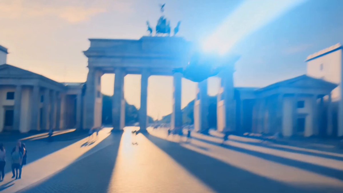 The image shows a large group of people walking around outside of the Brandenburg Gate in Berlin, Germany. The sun is shining brightly over the scene, creating long shadows that add depth and dimension to the photograph. The perspective of the photo suggests that it was taken from a distance or at an angle, capturing the grandeur of the monument as well as the bustling activity of people around it.