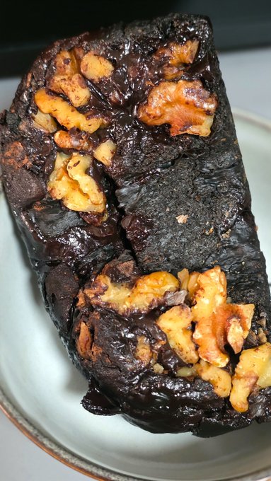 A rich, dark chocolate brownie or dessert studded with walnut pieces, served on a light-colored plate.