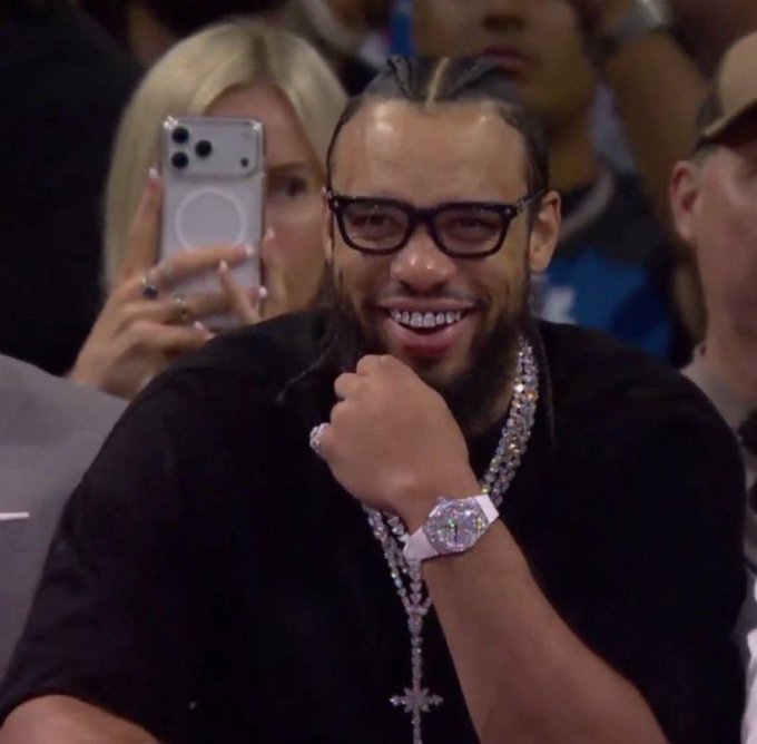 The image shows a smiling man with braided hair, glasses, a large diamond-encrusted watch, and a cross necklace being photographed in a crowd. The caption doesn't directly relate to the image's content, but rather uses the celebratory, triumphant expression as a metaphor for the author's satisfaction in watching the "pump" outperform the "fomo" platform during high-volume events like a Trump coin launch.