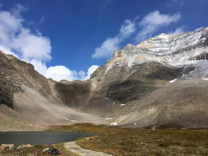 A scenic mountain landscape featuring a rocky, snow-dusted peak under a blue sky with scattered clouds, overlooking a small alpine lake with hikers resting near a trail in the foreground.